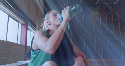 Female Athlete Drinking Water on Bench in Gymnasium During Workout Break with Sunbeams
