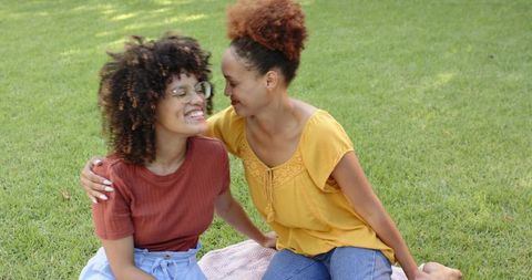 Two women laughing and embracing on picnic blanket in sunlit green park, candid couple