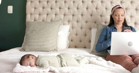 Mother Multitasking with Infant Sleeping Nearby on Bed