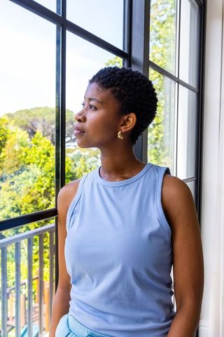 Contemplative african woman standing by window looking out at sunlit trees wearing light blue top