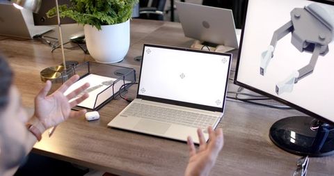 Indian man gesturing at laptop with robot gripper on monitor in modern coworking workspace