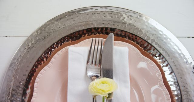 This elegant table setting includes a hammered metal charger, pink plates, and a delicate white flower atop a white napkin with fork and knife. Ideal for use in restaurant menus, wedding décor ideas, dining event invitations, or lifestyle blog posts related to dining etiquette and home décor.