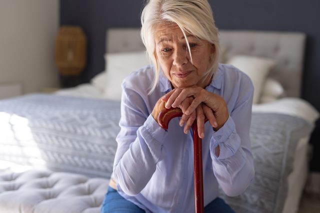 Thoughtful, mature caucasian woman sitting bedroom leaning on walking stick, copy space. Domestic life, living alone, health and senior lifestyle concept.