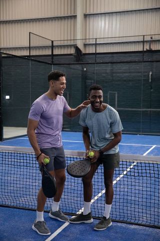 Diverse male friends laughing and holding paddles at indoor padel court net