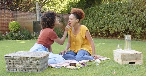 Midadult african american women sharing strawberry during sunlit backyard picnic, relaxing