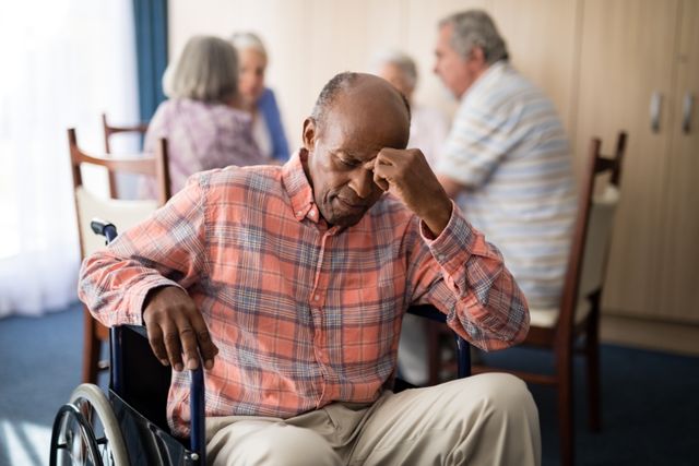 Elderly man sitting in a wheelchair, looking distressed and lonely in a retirement home. Other seniors are seen in the background, engaged in conversation. This image can be used to depict themes of aging, mental health, loneliness, and the challenges faced by seniors in assisted living facilities. Suitable for articles on elderly care, mental health awareness, and the importance of emotional support for seniors.