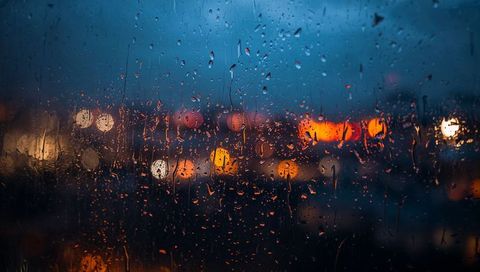 Raindrops streaking on glass window with city lights bokeh at dusk