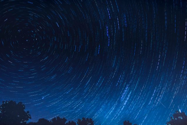 Star trails illuminating night sky above trees on forested horizon. Perfect for illustrating night photography, astronomy projects, science presentations, and nature-related content. Ideal for creating posters, backgrounds, wallpapers, and educational materials.