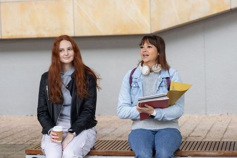 Female students sitting on campus bench holding coffee and folders, chatting casually