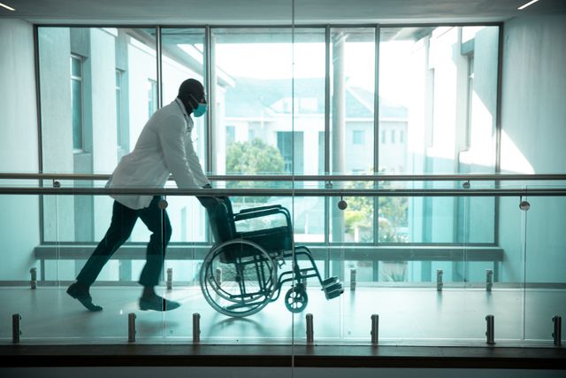 This image depicts an African American male doctor wearing a face mask and pushing an empty wheelchair in a hospital corridor. It highlights the importance of safety and healthcare services during the COVID-19 pandemic. This image can be used in articles, blogs, and websites related to healthcare, medical services, pandemic safety measures, and hospital environments.