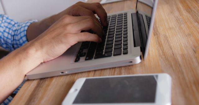 Hands typing on a laptop with a smartphone resting on a wooden table. Useful for illustrating remote work, office productivity, or technology usage in modern work environments.