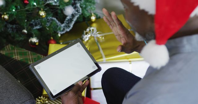 The image depicts a man wearing a Santa hat waving while holding a tablet in front of a decorated Christmas tree with gifts. It is ideal for illustrating concepts related to virtual celebrations, remote holidays, and staying connected with loved ones during the holiday season. It can be used for articles, advertisements, or social media posts about celebrating holidays remotely and utilizing modern technology.