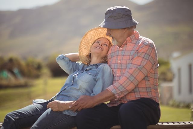 Senior couple enjoying a sunny day in the countryside, sitting on a bench and smiling at each other. Ideal for use in advertisements or articles about retirement, healthy aging, outdoor activities, and senior lifestyle. Perfect for promoting products or services related to elderly care, travel, and leisure.