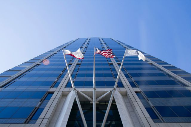 Flags from several countries wave on flagpoles positioned in front of a modern high-rise building with a sleek glass facade. The clear blue sky and urban setting create a polished and professional atmosphere, ideal for use in articles about international business, corporate outfits, globalization, urban development, and modern architecture. It can also be used to represent corporate offices or patriotic themes.