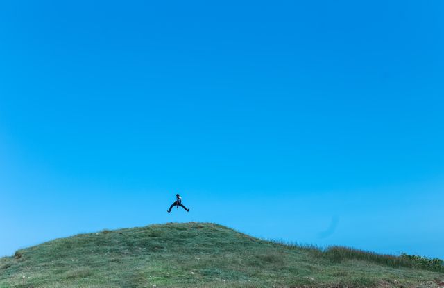 Person leaping on a serene grassy hilltop under a cloudless blue sky. Ideal for depicting outdoor activities, freedom, adventure, joy, and connection with nature. Perfect for travel brochures, wellness blogs, motivational posters, and outdoor adventure campaigns.