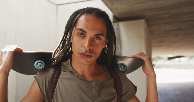 Portrait of fashionable biracial man with dreadlocks holding skateboard and looking at camera. Street style and modern urban lifestyle.
