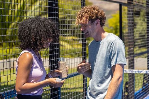 Laughing diverse young couple sharing reusable water bottle on sunny outdoor sports court
