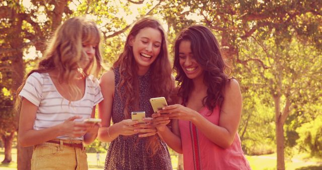This image captures three teenage girls enjoying each other's company while using their smartphones in a sunny park. They are laughing and appear to be sharing something on their devices. This image can be used in articles or advertisements related to teenage social interactions, friendship, the impact of technology on youth, or lifestyle content emphasizing outdoor activities.