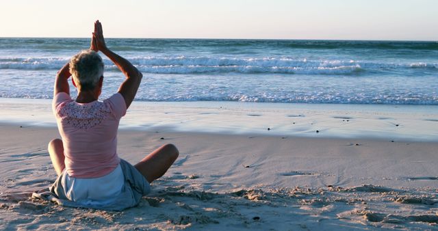 This image shows a senior woman seated on the sand with hands clasped overhead in a yoga pose, practicing meditation and relaxation on a beach during sunset. Perfect for use in articles or advertisements related to mental well-being, senior lifestyle, wellness retreats, and travel. Useful for promotional materials for yoga classes, meditation sessions, and health-focused content.