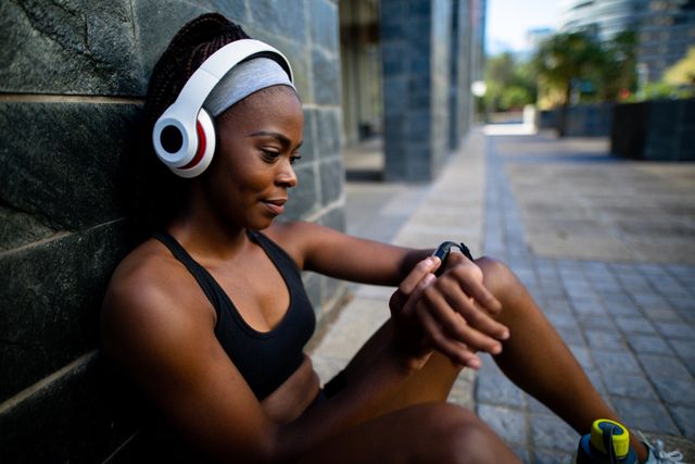 African American woman sitting against a wall, wearing headphones and sportswear, checking her smartwatch. Ideal for promoting fitness, healthy lifestyle, wearable technology, urban exercise routines, and sports gear.
