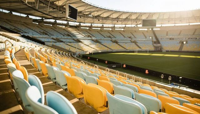 Empty soccer stadium curved yellow light blue seats sunlit pitch staff broadcast screens