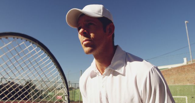 Young male tennis player wearing white clothing and cap, standing on outdoor tennis court with focused expression. Ideal for use in sports events promotion, athletic apparel advertisements, motivational sports posters.