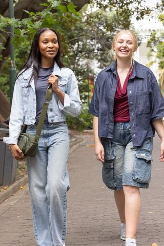 Two friends walking on park path wearing denim and carrying olive crossbody bag, smiling