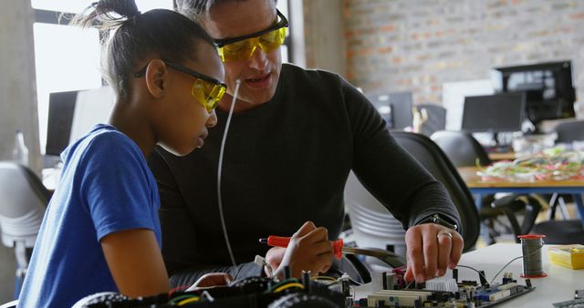 Mentor guiding young student in robotics class, providing hands-on learning experience in a STEM environment. Wearing safety glasses, they are working on an electronics circuit board, indicating an emphasis on safety and practicality. This can be used for materials promoting educational programs in science and technology, articles on mentorship in STEM, or advertisements for tech workshops.