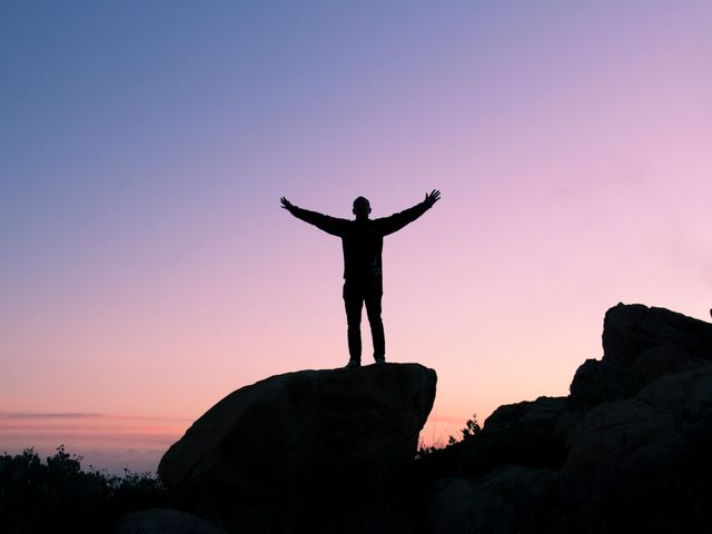 A person stands on a rock with open arms, silhouetted against the evening sky at dusk. This image conveys a sense of freedom and triumph, making it ideal for use in motivational or inspirational contexts, adventure and travel promotions, and outdoor activity advertisements.