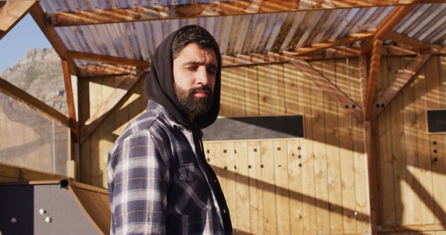 Man wearing hoodie and plaid shirt standing outdoors in rustic environment with wooden structure and sunlight casting shadows. Ideal for themes of outdoor lifestyle, fashion, rugged individualism, natural settings.