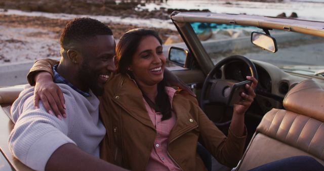 Happy couple enjoying time by the beach in a convertible car at sunset, taking a selfie and sharing a moment of joy. Ideal for themes of romance, vacation, carefree lifestyle, travel adventures, and memorable experiences.