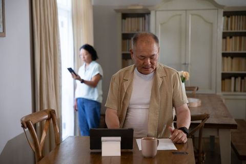 Asian seniors using tablet and smartphone at dining table in sunlit minimalist home