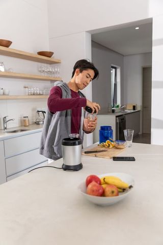 Asian man preparing protein smoothie in modern minimalist kitchen with blender and fruit