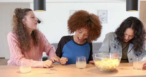 Diverse friends laughing, cheering and sharing popcorn at bright modern kitchen table