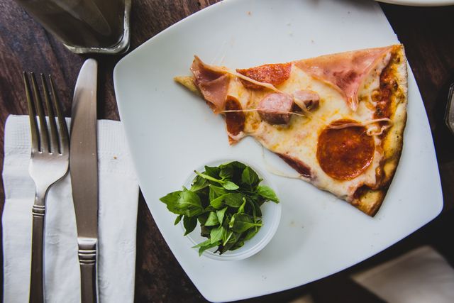 A delicious pizza slice with pepperoni and ham is placed on a white plate. Beside it, fresh herbs in a small cup add a touch of greenery. The cutlery is neatly placed on a folded napkin, resting on a wooden table. Ideal for use in food blogs, restaurant promos, Italian cuisine features, and culinary magazine articles.