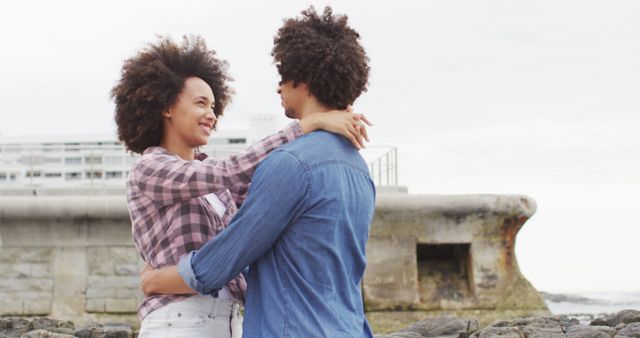 Young couple with afro hair embracing outdoors, wearing casual clothes on a cloudy day. The woman is smiling while looking at her partner. Use for content related to relationships, love, romance, happiness, and lifestyle themes.