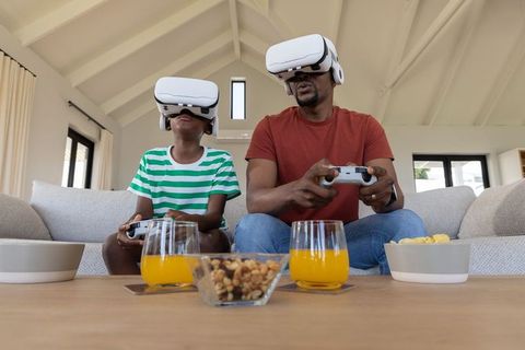 Father and son playing vr games on living room sofa wearing vr headsets using controllers