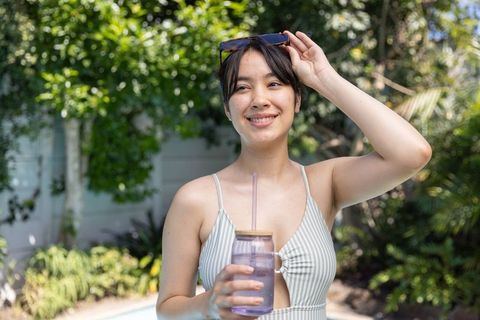 Smiling asian woman holding tumbler by pool lifting sunglasses in striped swimsuit