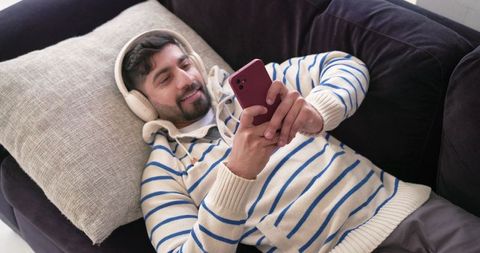 Indian man reclining on sofa wearing striped sweater and headphones using smartphone