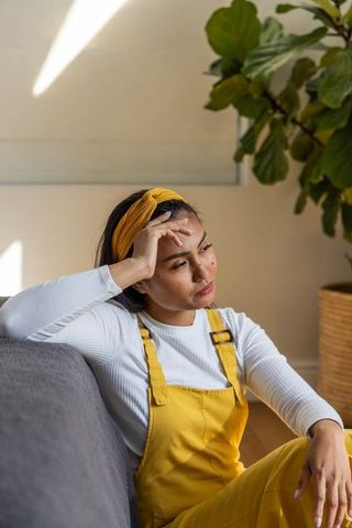 Pensive woman in mustard overalls and headband sitting on gray sofa beside indoor plant