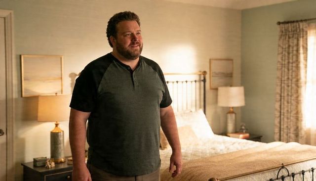 Middle-aged man standing in sunlit bedroom with metal headboard and bedside lamps