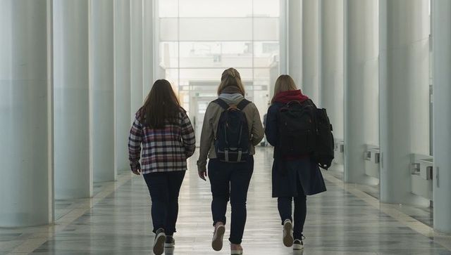 Three Students Walking Down Bright Campus Corridor With Backpacks and Coats