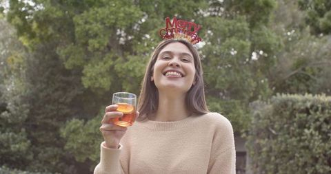 Smiling woman wearing merry christmas headband holding glass of festive drink in garden