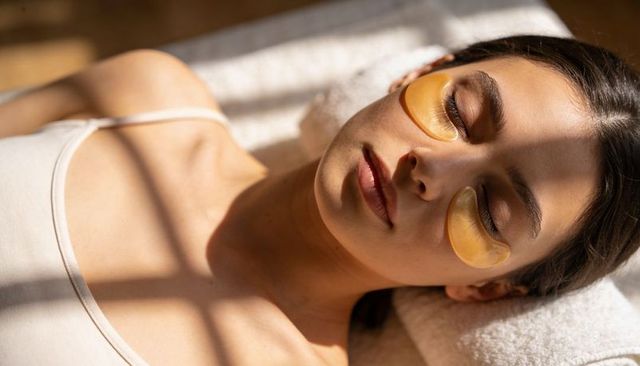 Sunlit woman relaxing with gold under-eye hydrogel patches on spa bed rejuvenating skin