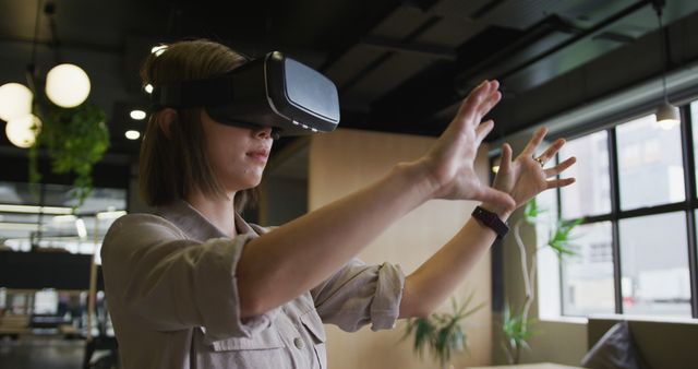 Woman using virtual reality headset indoors with arms outstretched, showcasing advanced technology in modern office environment. Perfect for illustrating concepts of innovation, digital interaction, augmented reality, and cutting-edge technology in professional or educational settings.