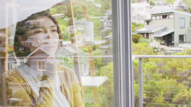 This video depicts an Asian woman standing by a window, touching her chin with a thoughtful expression. It is ideal for use in themes of introspection, solitude, peaceful moments at home, lifestyle articles, and mental well-being. The reflection in the window adds depth, making it suitable for visual storytelling about personal thoughts, decisions, and indoor settings.