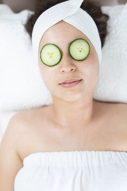Caucasian woman lying in a beauty salon with cucumber slices on her eyes, enjoying a facial treatment. Ideal for use in articles or advertisements related to skincare, beauty treatments, spa services, wellness, and self-care routines. Perfect for illustrating relaxation and pampering in a professional beauty setting.