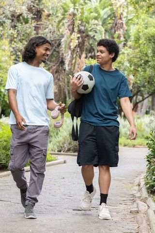 African american young men walking in park holding soccer ball and headphones conversing