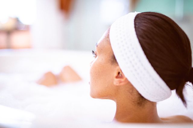 Biracial woman enjoying a relaxing foam bath at home, wearing a white headband. Perfect for themes related to self care, wellness, and domestic lifestyle. Ideal for use in articles, blogs, and advertisements promoting relaxation, spa treatments, and personal care products.
