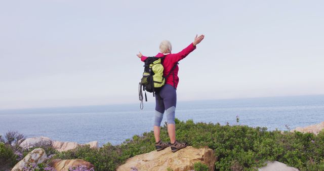 Rear view of senior hiker woman standing with her arms wide open standing on a rock while trekking in the mountains. trekking, hiking, nature, activity, exploration, adventure concept.